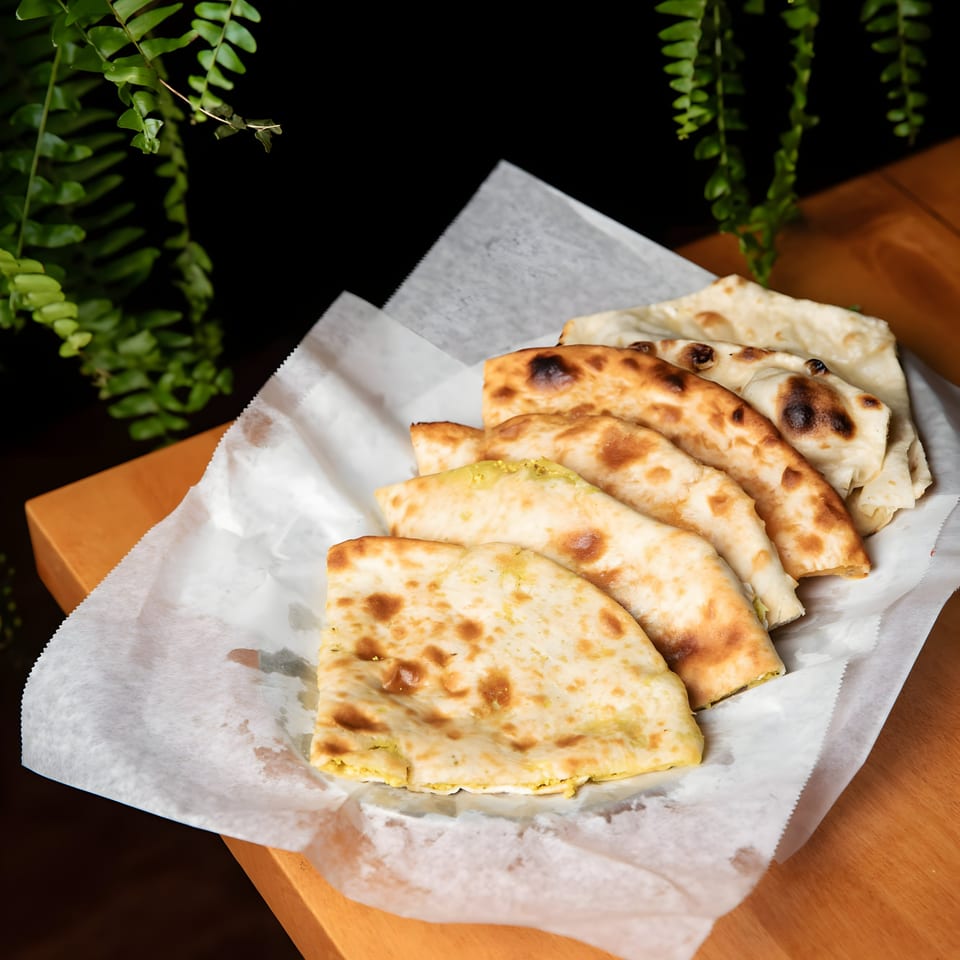 A basket of freshly baked Indian naan bread, some plain, some stuffed, on a wooden table.