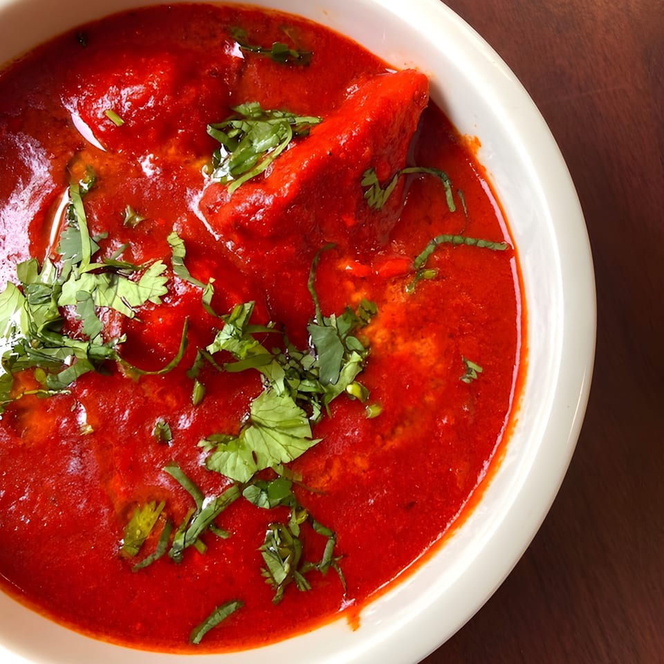 Close-up of vibrant red Indian curry with chicken pieces and fresh green cilantro in a white bowl.